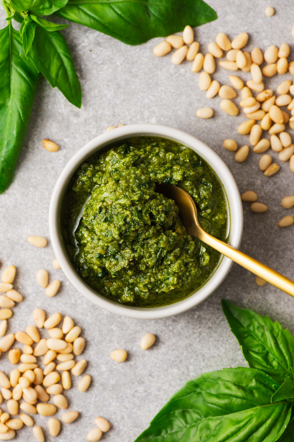 Traditional Italian basil pesto in a white bowl surrounded by typical pesto ingredients including pine nuts and Genovese basil.