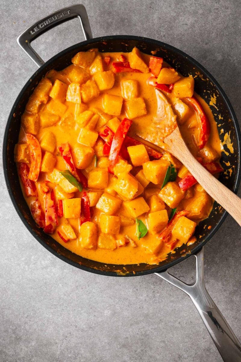 Cubed butternut being cooked in red curry sauce with red bell pepper and lime leaves, stirred with a wooden spoon.